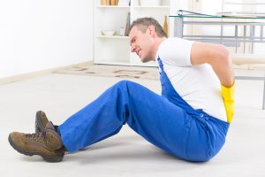 A worker in blue overalls sat on the floor holding his lower back in pain after a fall at work.